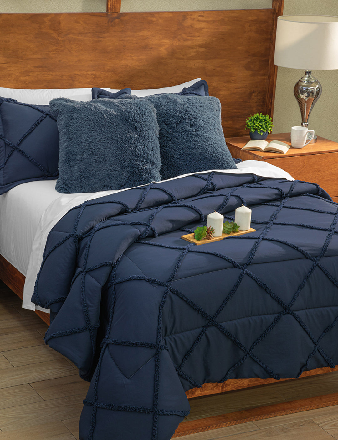 A neatly made bed with Intima Hogar’s Coordinado de Edredón Luxury Seúl in navy blue diamond pattern, two fuzzy navy pillows, white Royal Cotton sheet set, a wooden headboard, nightstand with lamp, plant, and candles on a tray.