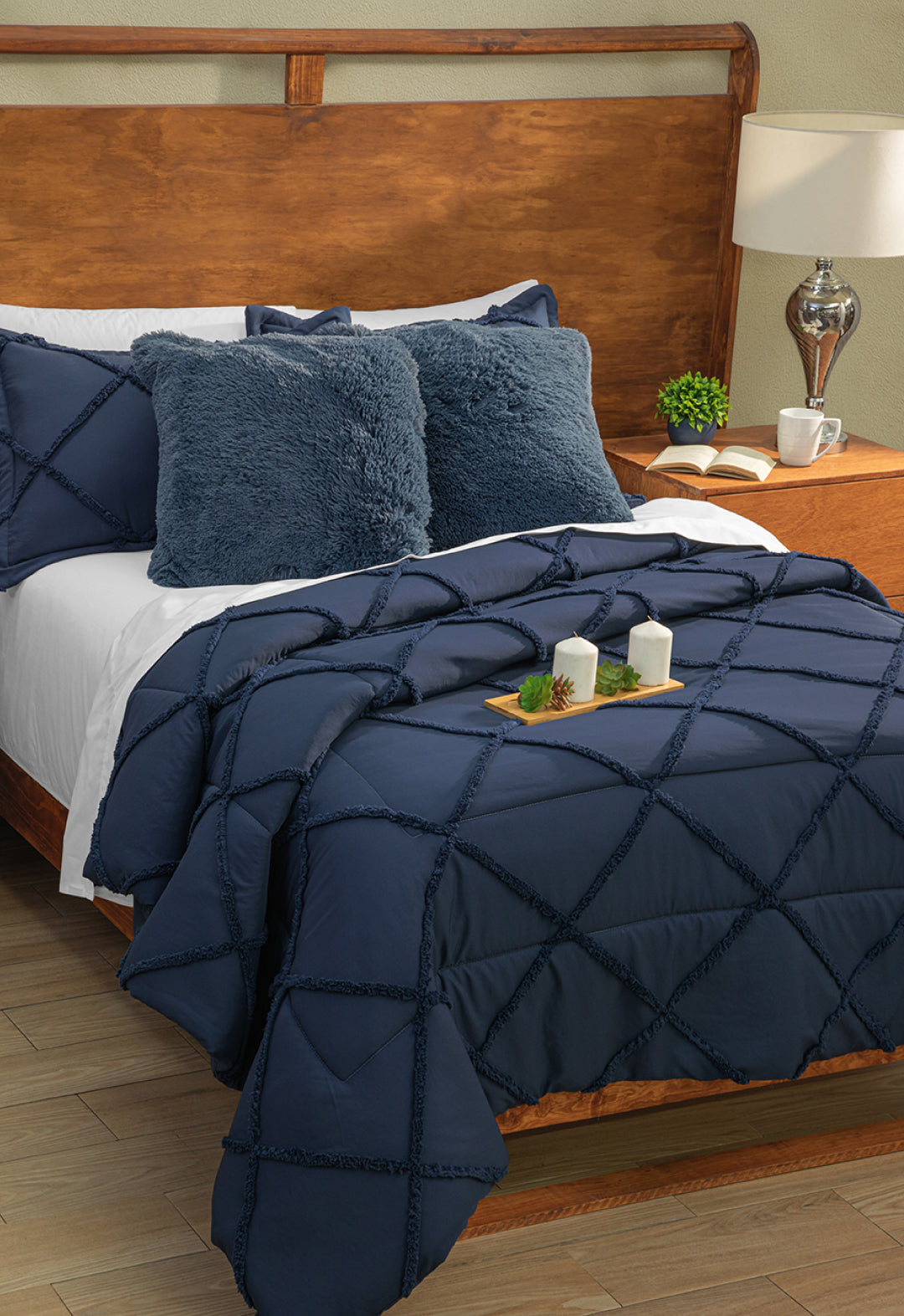 A neatly made bed with Intima Hogar’s Coordinado de Edredón Luxury Seúl in navy blue diamond pattern, two fuzzy navy pillows, white Royal Cotton sheet set, a wooden headboard, nightstand with lamp, plant, and candles on a tray.