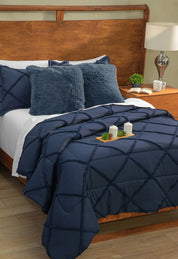 A neatly made bed with Intima Hogar’s Coordinado de Edredón Luxury Seúl in navy blue diamond pattern, two fuzzy navy pillows, white Royal Cotton sheet set, a wooden headboard, nightstand with lamp, plant, and candles on a tray.