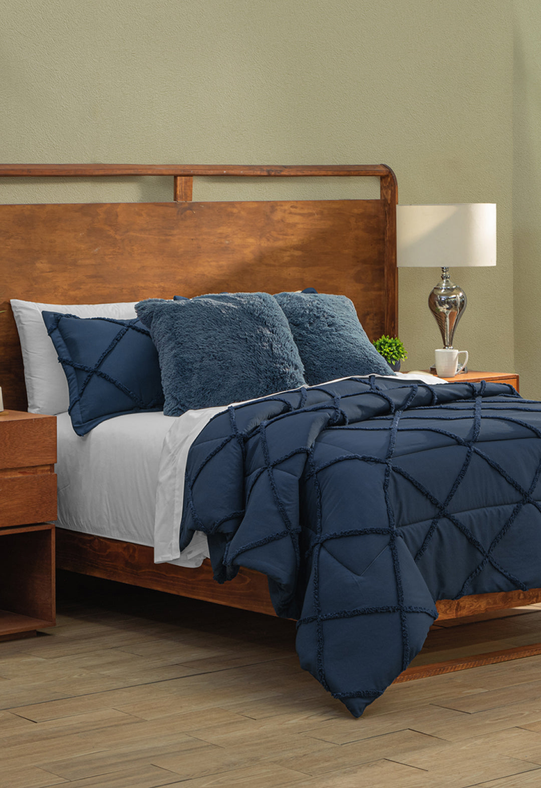 A wooden bed features the Coordinado de Edredón Luxury Seúl by Intima Hogar, paired with white Royal Cotton sheets and pillows. A bedside table holds a lamp, cup, and small plant in a light-walled room with wood floors.