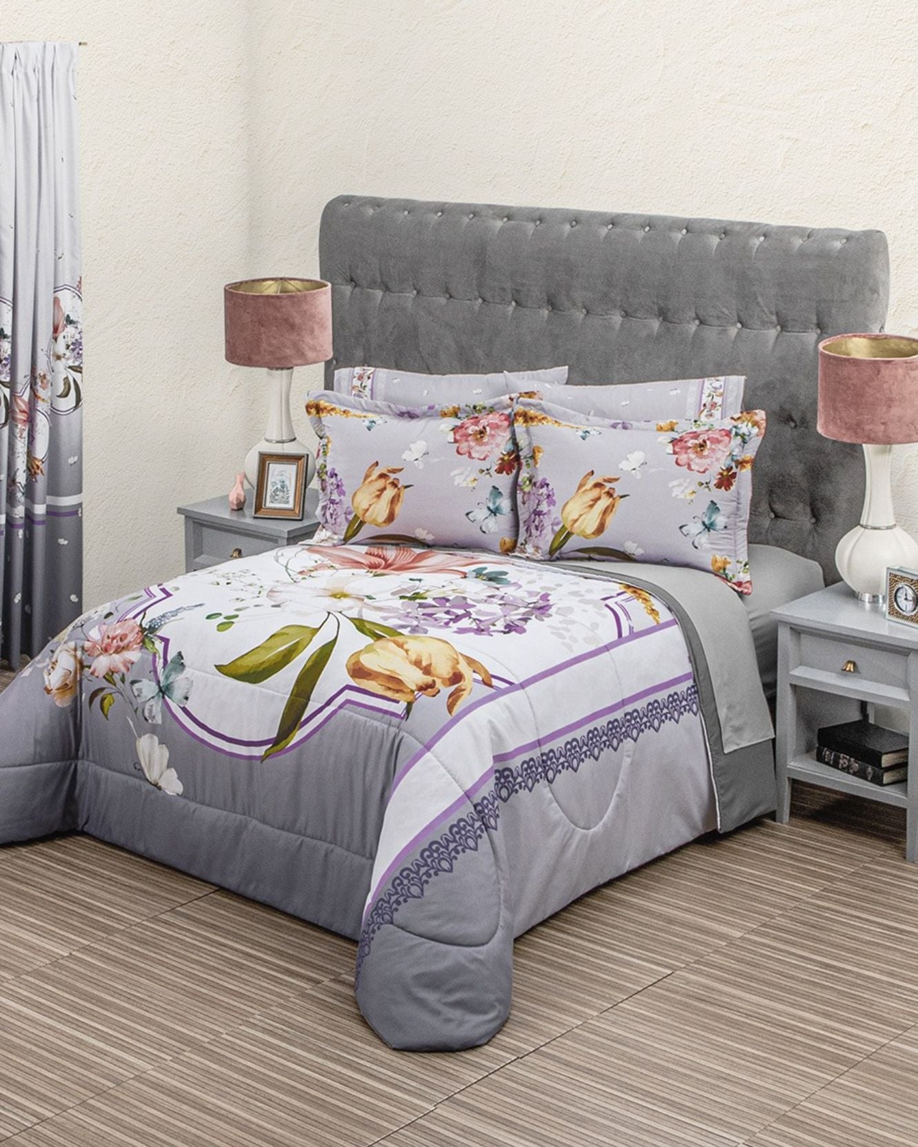 A neatly made bed with the Coordinado de Edredón Super Suave Susan by Intima Hogar in gray, purple, and beige floral tones, paired with a tufted headboard, two nightstands with white lamps, and a framed photo against a light wall.
