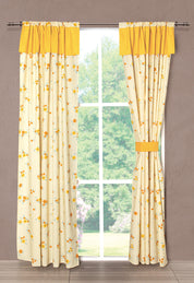 A window adorned with the Juego de Cortinas Girasoles by Intima Hogar features light beige curtains with a yellow floral pattern and solid yellow valances, tied back on one side, allowing natural light and a view of greenery.