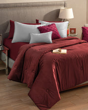 A neatly made bed features Intima Hogar’s Funda de Almohada Ensueño Gris, burgundy comforter, matching pillows, and decorative cushions. A small gift box rests on the bed beside a lamp, clock, and book for your descanso.