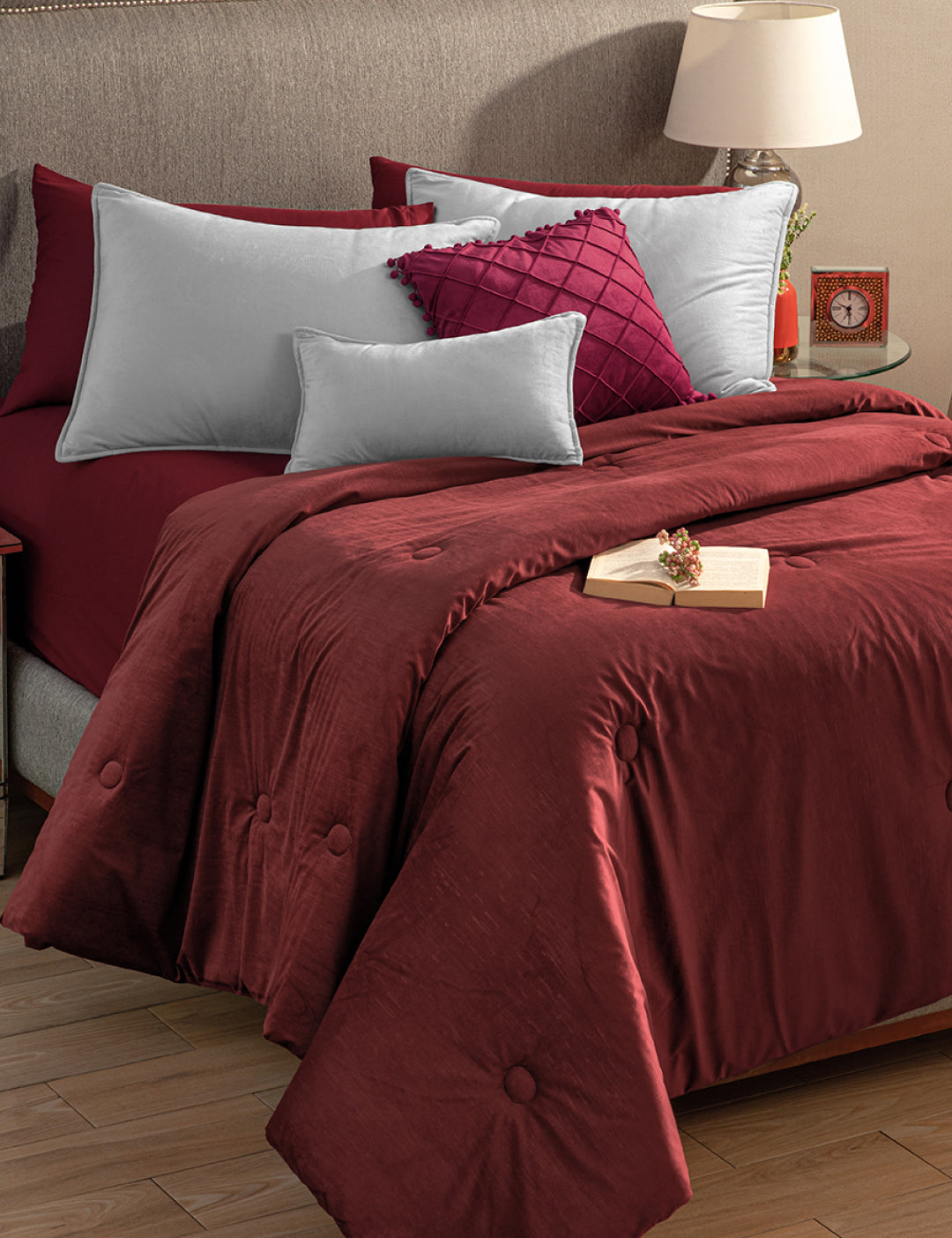 A neatly made bed features the Edredón Súper Suave Janeiro by Intima Hogar in burgundy, silver-gray and burgundy pillows, a taupe upholstered headboard, and a wooden nightstand with a lamp, clock, decor, and an open book.