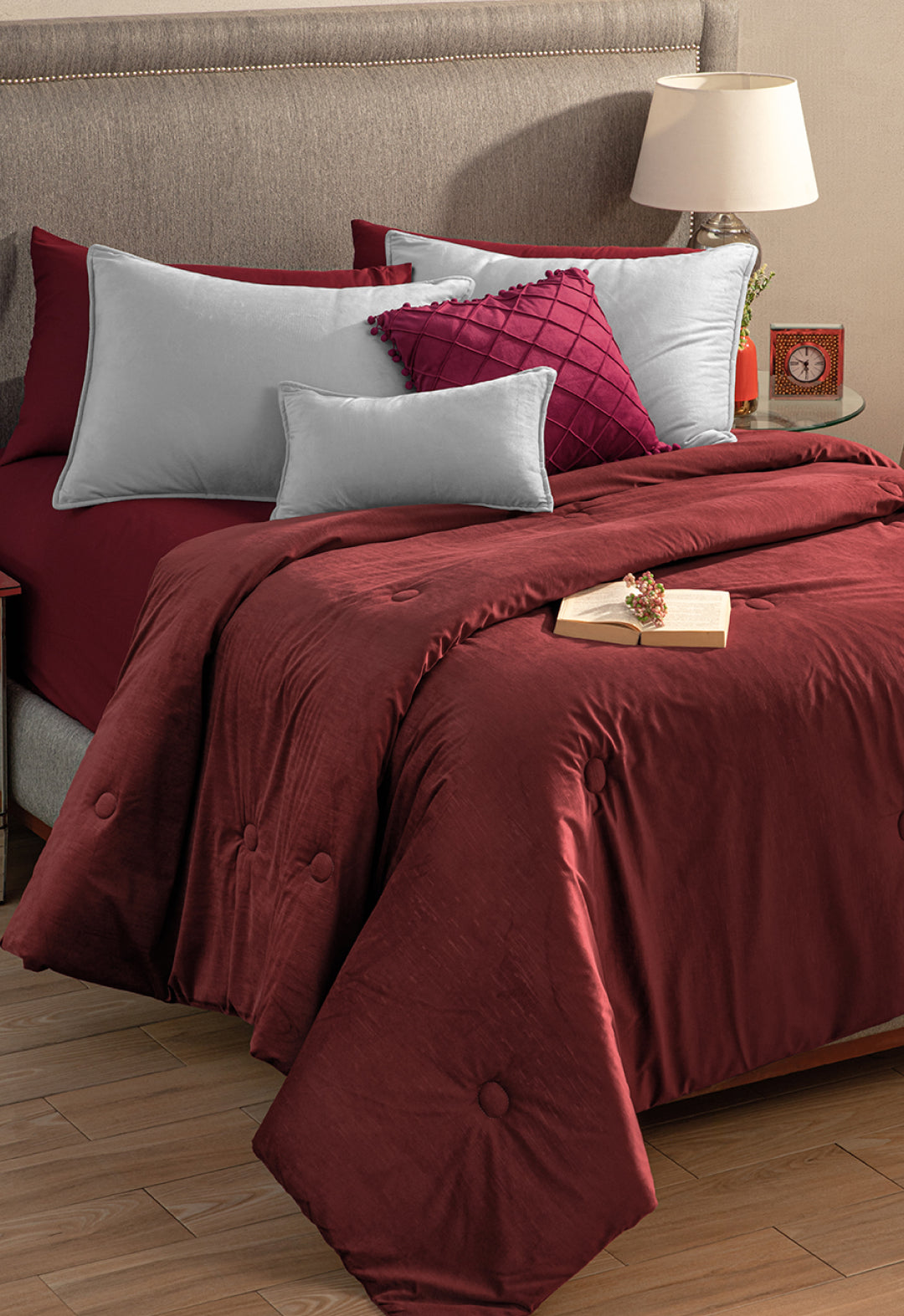 A neatly made bed features the Edredón Súper Suave Janeiro by Intima Hogar in burgundy, silver-gray and burgundy pillows, a taupe upholstered headboard, and a wooden nightstand with a lamp, clock, decor, and an open book.