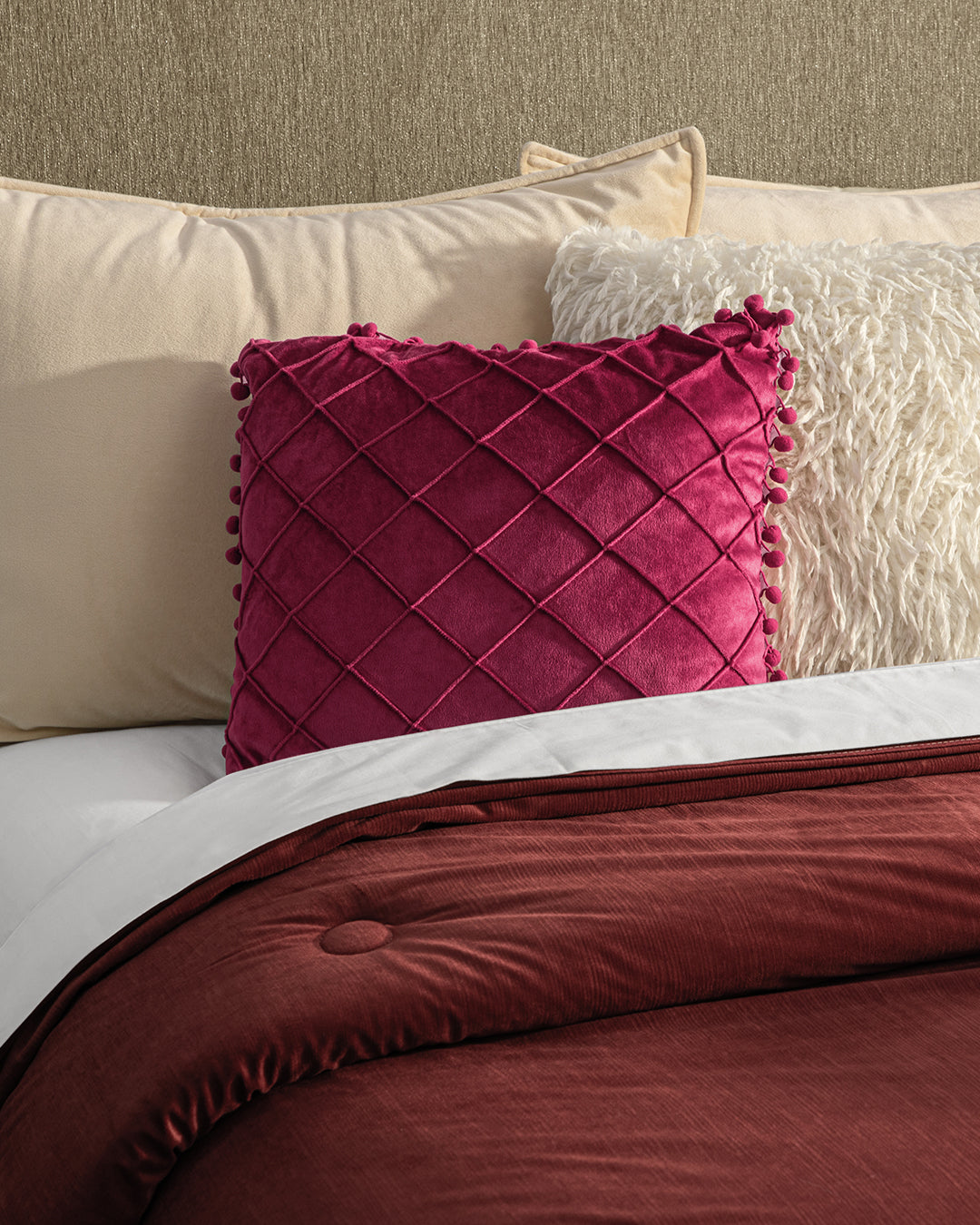 A neatly made bed with a burgundy comforter, beige pillows, a textured white pillow, and the Cojín de Lujo Velvet Vino from Intima Hogar—a plush velvet accent with quilted details—adds lujo sutil to the center.