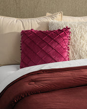 A neatly made bed with a burgundy comforter, beige pillows, a textured white pillow, and the Cojín de Lujo Velvet Vino from Intima Hogar—a plush velvet accent with quilted details—adds lujo sutil to the center.