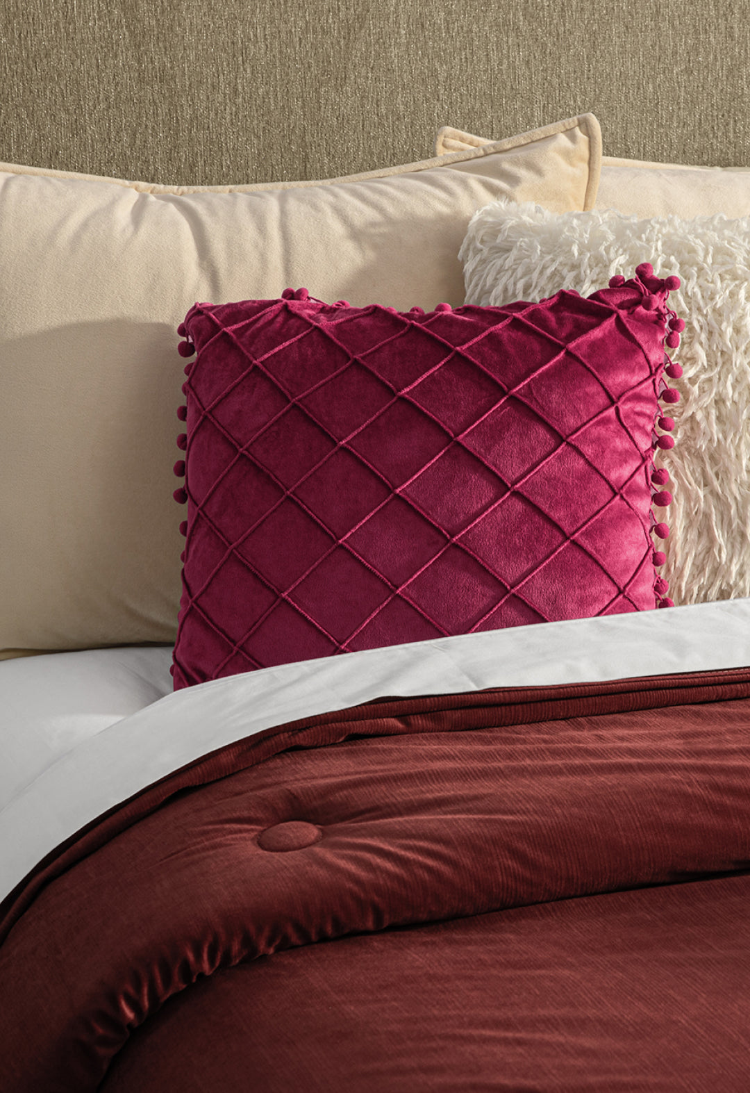 A neatly made bed features cream and off-white pillows, a textured cream pillow, a magenta diamond-patterned decorative pillow, and the Edredón Súper Suave Janeiro by Intima Hogar in rich burgundy.