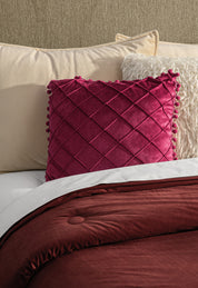 A neatly made bed features cream and off-white pillows, a textured cream pillow, a magenta diamond-patterned decorative pillow, and the Edredón Súper Suave Janeiro by Intima Hogar in rich burgundy.