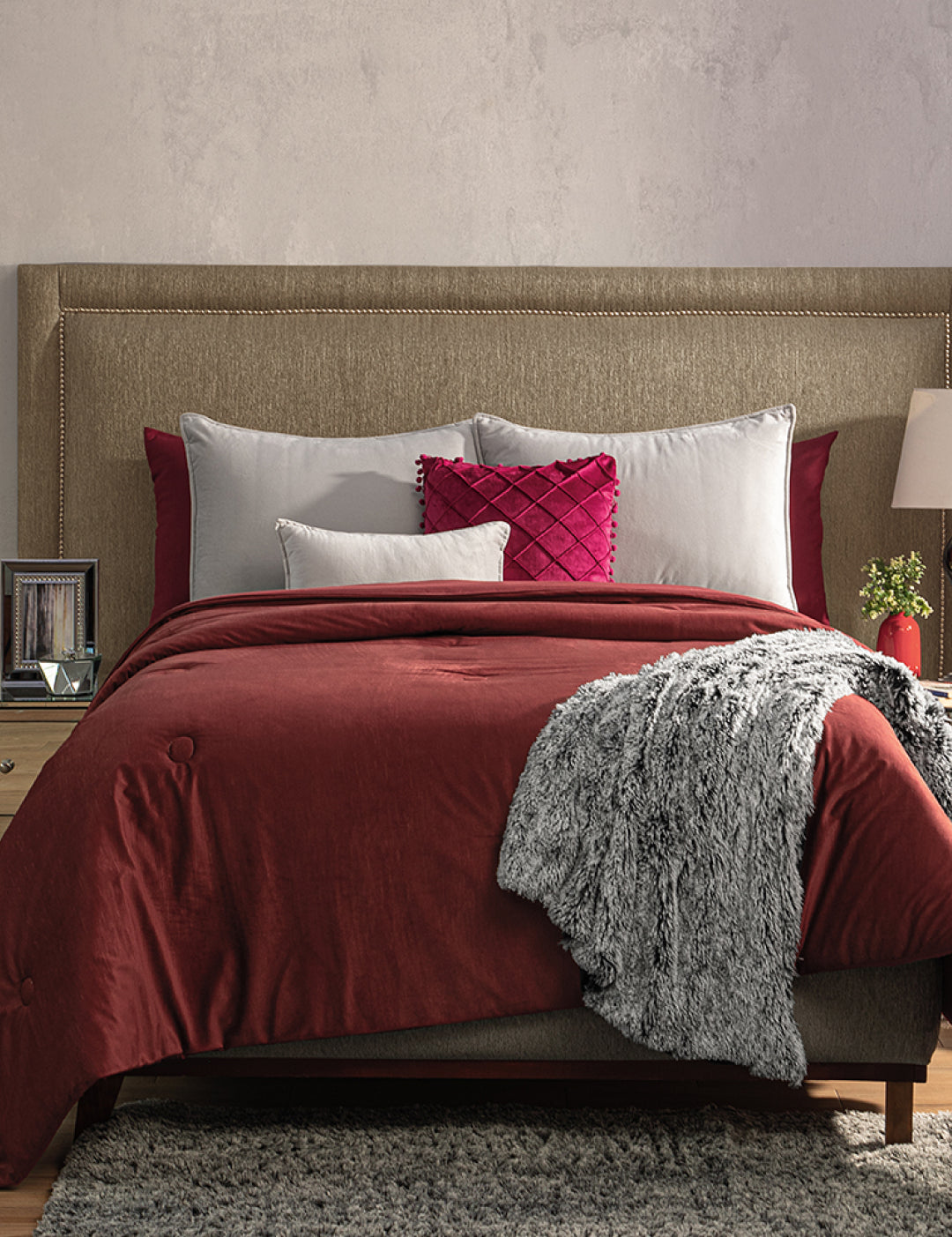 A neatly made bed features the Edredón Súper Suave Janeiro by Intima Hogar in red, gray and white pillows, a textured gray throw blanket, and a beige upholstered headboard. Nightstands with lamps and decor are on each side.
