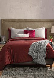 A neatly made bed features the Edredón Súper Suave Janeiro by Intima Hogar in red, gray and white pillows, a textured gray throw blanket, and a beige upholstered headboard. Nightstands with lamps and decor are on each side.