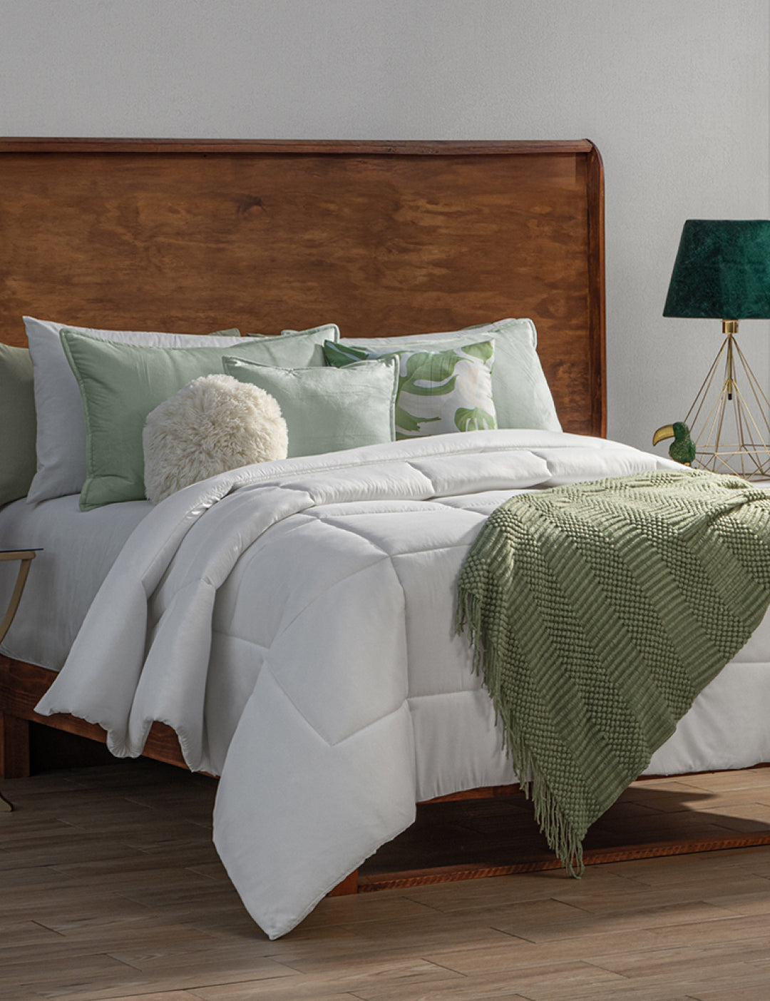 A neatly made bed with white bedding features the Edredón Súper Suave Maxiplus by Intima Hogar for calidez envolvente. Light green pillows, a fluffy round cushion, and a green lamp on a gold base add comfort next to the wooden headboard.