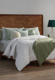 A neatly made bed with white bedding features the Edredón Súper Suave Maxiplus by Intima Hogar for calidez envolvente. Light green pillows, a fluffy round cushion, and a green lamp on a gold base add comfort next to the wooden headboard.
