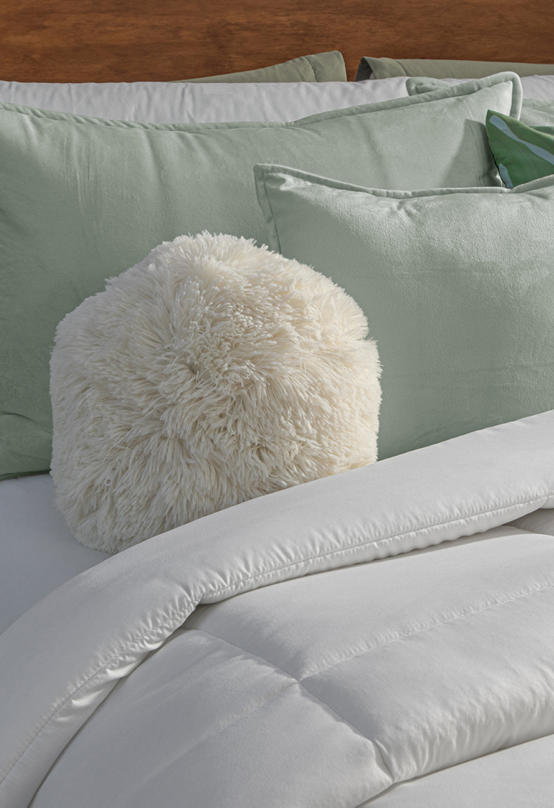 A white fluffy round pillow rests on a bed with sage green pillows and the Edredón Súper Suave Maxiplus by Intima Hogar, creating a cozy and inviting atmosphere of calidez envolvente.