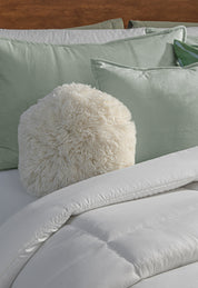 A white fluffy round pillow rests on a bed with sage green pillows and the Edredón Súper Suave Maxiplus by Intima Hogar, creating a cozy and inviting atmosphere of calidez envolvente.