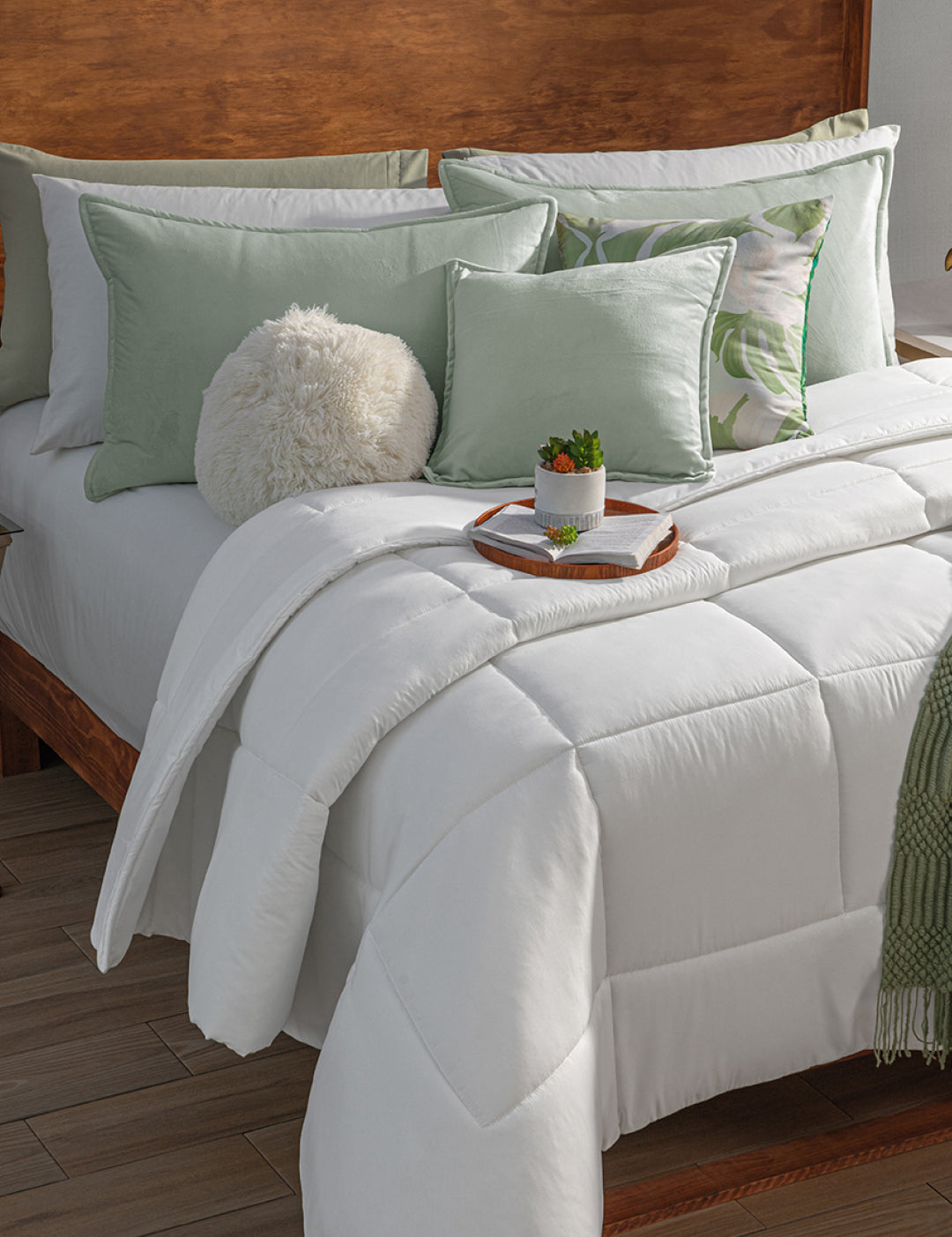 A neatly made bed features the Edredón Súper Suave Maxiplus by Intima Hogar, green and white accent pillows, a fluffy round pillow, and a tray with a plant and cup—all set against a wooden headboard and warm brown flooring for calidez envolvente.