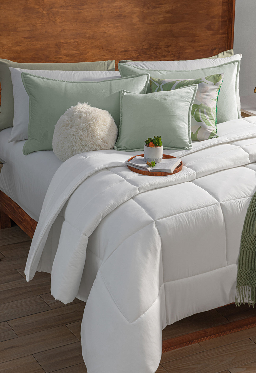 A neatly made bed features the Edredón Súper Suave Maxiplus by Intima Hogar, green and white accent pillows, a fluffy round pillow, and a tray with a plant and cup—all set against a wooden headboard and warm brown flooring for calidez envolvente.
