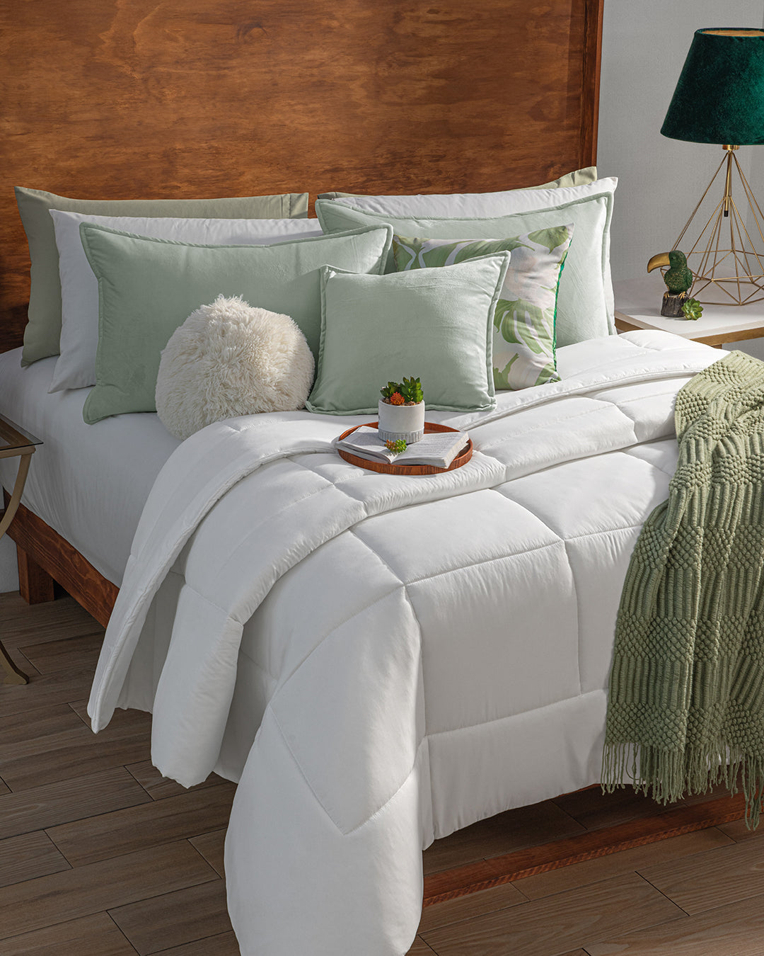 A neatly made bed with Intima Hogar's Funda de Almohada Ensueño Verde Pistacho, white bedding, a green throw, and a decorative tray. The wooden headboard and modern nightstand create a cozy space ideal for relaxing.