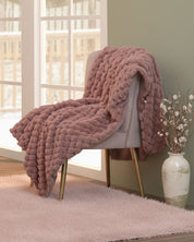 La Frazada de Lujo Aruba de Intima Hogar, en un cálido tono palo rosa, está sobre una silla beige moderna con patas doradas junto a un jarrón decorativo, aportando elegancia y calidez a esta acogedora habitación con alfombra rosa suave.