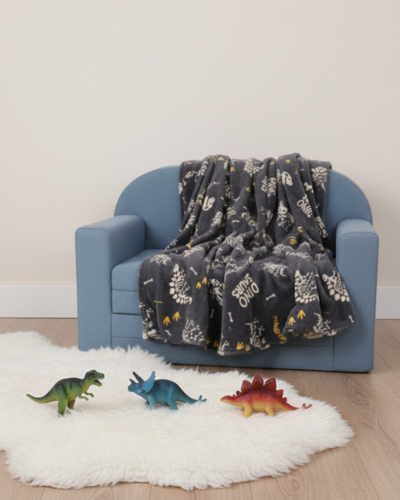 A small blue sofa is draped with the Intima Hogar Frazada Bones, featuring a dinosaur pattern. On the wooden floor, a white fluffy rug holds three toy dinosaurs: T. rex, triceratops, and stegosaurus.
