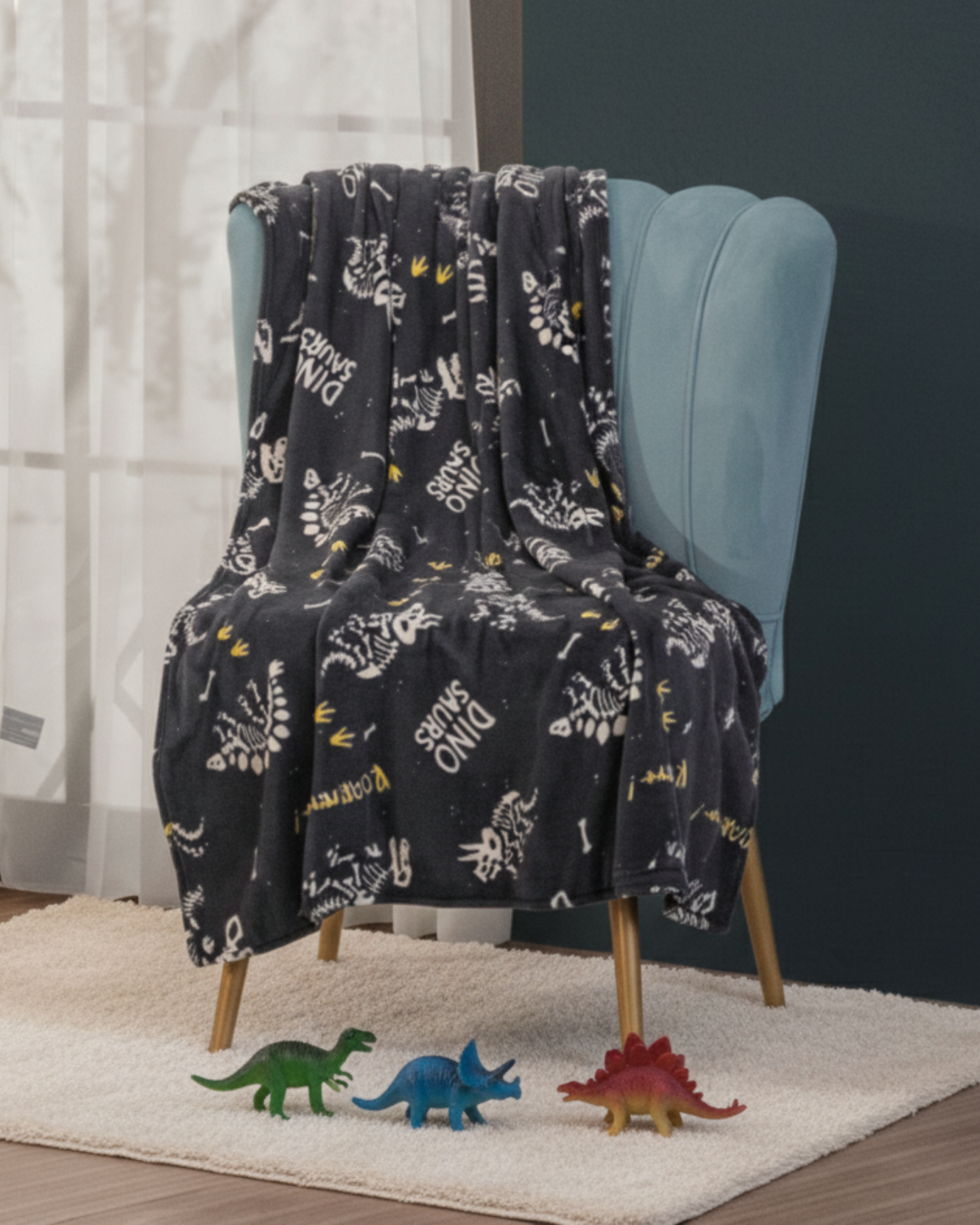 The Frazada Bones by Intima Hogar, featuring dark dinosaur skeletons and text, is draped over a light blue chair. Three small toy dinosaurs sit on a white rug, with a window and a dark green wall in the background.