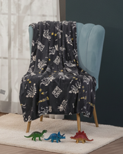 The Frazada Bones by Intima Hogar, featuring dark dinosaur skeletons and text, is draped over a light blue chair. Three small toy dinosaurs sit on a white rug, with a window and a dark green wall in the background.