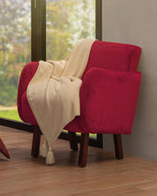 A red velvet armchair with wooden legs sits by a large window. Draped over one arm is the Frazada de Lujo Chai Beige by Intima Hogar. The wooden floor gleams, and trees are visible outside.