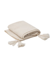 The Frazada de Lujo Chai Beige by Intima Hogar is a folded cream-colored throw with a textured pattern and large corner pompons, displayed on a white background.