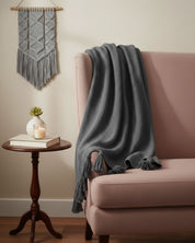 A pink sofa with the Frazada de Lujo Chai Gris Oxford by Intima Hogar draped over one arm sits beside a round wooden table holding a book, a small potted succulent, and a lit candle. A gray macramé wall hanging decorates the beige wall.