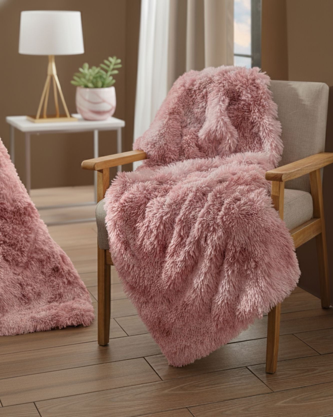 The Frazada de Lujo Flamenco by Intima Hogar, in soft pink, is draped over a beige cushioned wooden chair in a cozy modern room with wood floors. A small white table with a lamp and plant completes this elegant decorative scene.