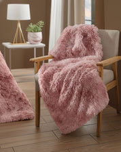 The Frazada de Lujo Flamenco by Intima Hogar, in soft pink, is draped over a beige cushioned wooden chair in a cozy modern room with wood floors. A small white table with a lamp and plant completes this elegant decorative scene.