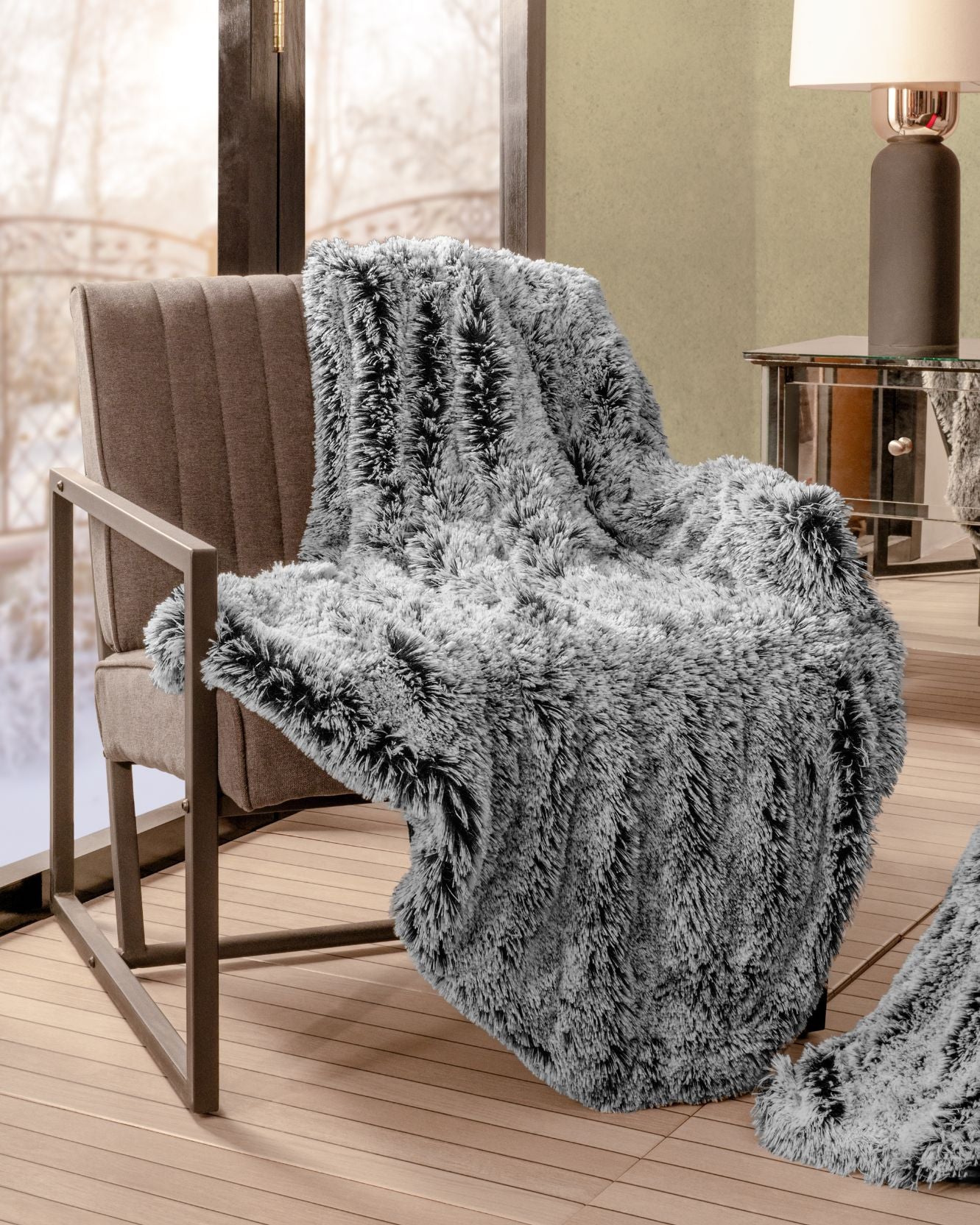 The Frazada de Lujo Humo by Intima Hogar is draped over a modern brown armchair beside a large window in a cozy, softly lit room with a side table and lamp, adding frescura y comodidad to the space.