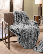 The Frazada de Lujo Humo by Intima Hogar is draped over a modern brown armchair beside a large window in a cozy, softly lit room with a side table and lamp, adding frescura y comodidad to the space.