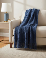 A cream-colored sofa with the Intima Hogar Frazada de Lujo Matcha Azul draped over the armrest, beside a wooden side table with a lamp and decor, brings coziness to this warmly lit room.