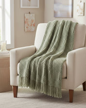 The Frazada de Lujo Matcha Verde by Intima Hogar, a soft sage green throw with fringe, is draped over a cream armchair in a bright, cozy living room with neutral décor and soft artwork.