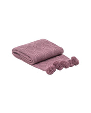 The Intima Hogar Frazada de Lujo Taro Lila is a folded mauve throw with a diamond pattern and pom-poms on the ends, shown on a white background.