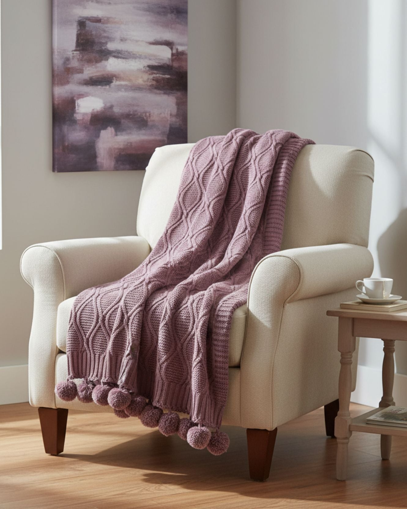 A cream-colored armchair is draped with the Intima Hogar Frazada de Lujo Taro Lila, featuring a cable pattern and pompons. A side table holds a white teacup and saucer, while abstract art hangs on the wall behind.