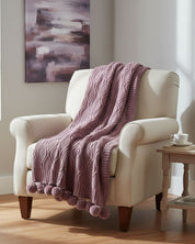 A cream-colored armchair is draped with the Intima Hogar Frazada de Lujo Taro Lila, featuring a cable pattern and pompons. A side table holds a white teacup and saucer, while abstract art hangs on the wall behind.