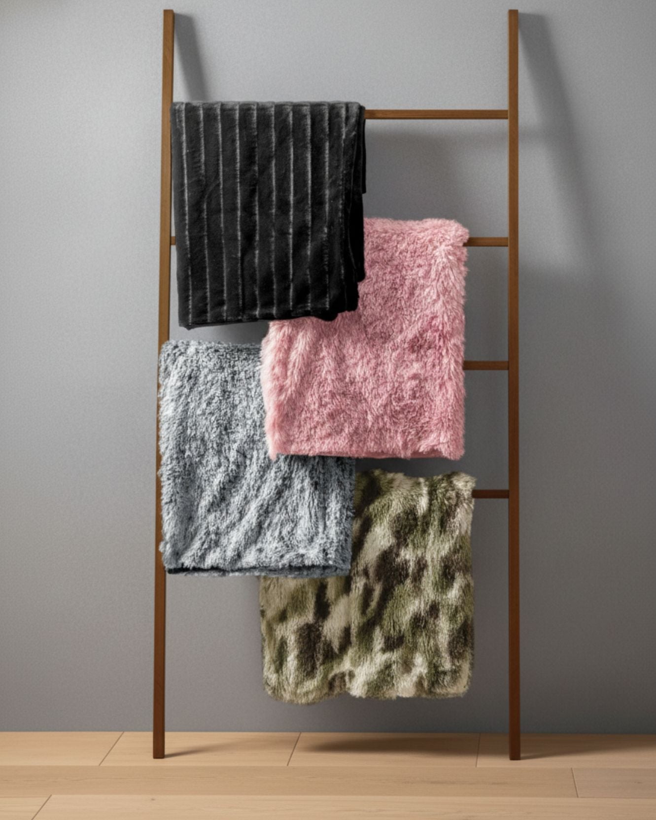 A brown wooden ladder rack from Intima Hogar leans against a gray wall, displaying four folded blankets—one is the Frazada de Lujo Humo—for added comfort and style. The rack stands on a light wooden floor.