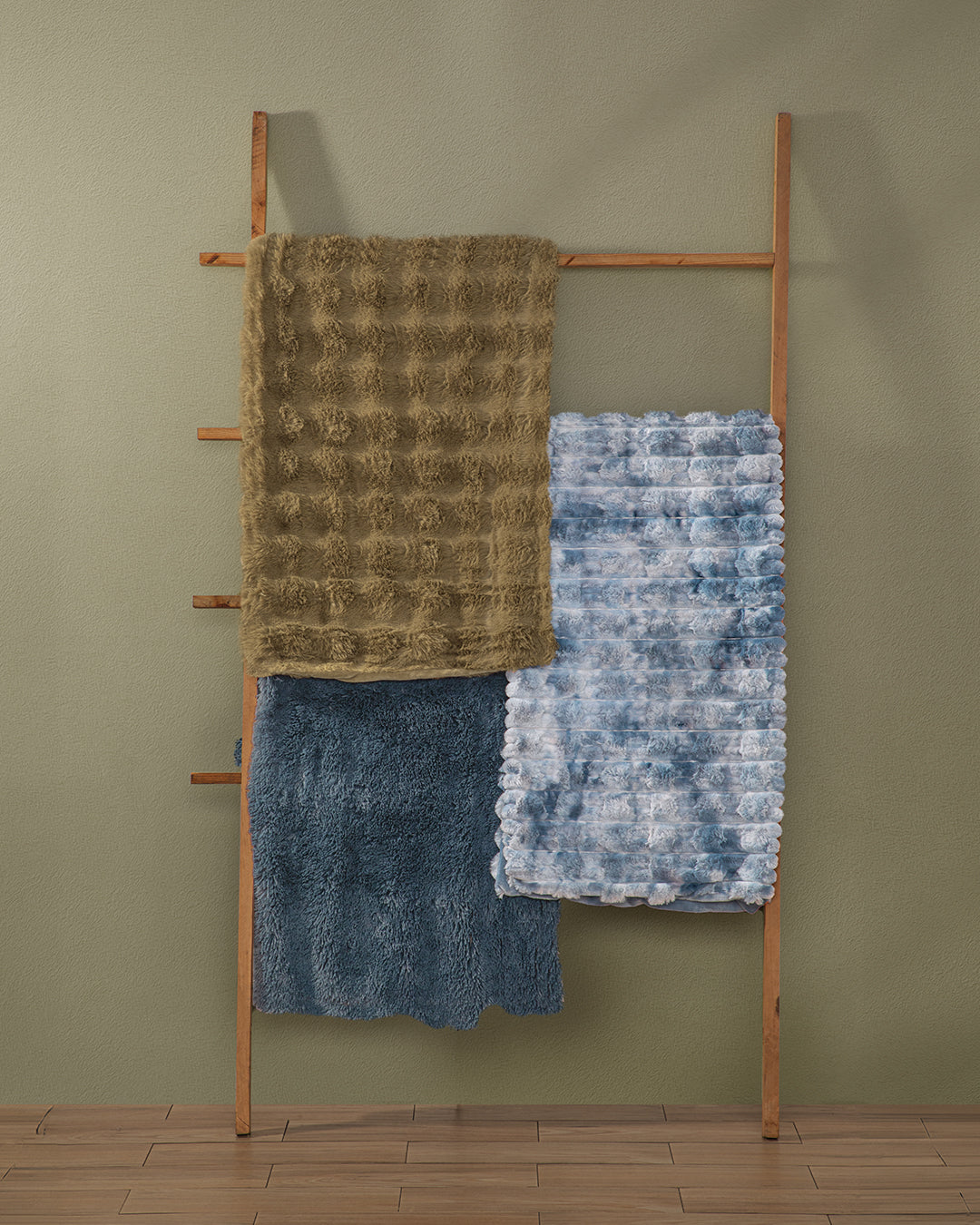 A wooden ladder leans against a green wall, displaying three textured blankets: an Intima Hogar Frazada de Lujo Quebec on the top rung, a blue tie-dye blanket in the middle, and a solid dark blue esponjosa blanket on the bottom. The floor is wooden.
