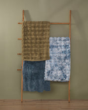 A wooden ladder leans against a green wall, displaying three textured blankets: an Intima Hogar Frazada de Lujo Quebec on the top rung, a blue tie-dye blanket in the middle, and a solid dark blue esponjosa blanket on the bottom. The floor is wooden.