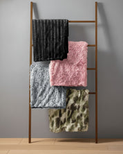 A wooden ladder against a gray wall displays four folded blankets, including the Intima Hogar Frazada de Lujo Flamenco in pink, a black quilted blanket, a fluffy gray frazada moderna, and one with a green camouflage pattern.