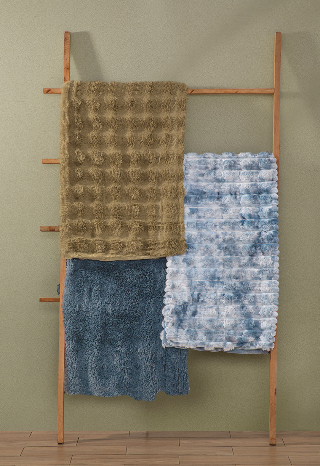 A wooden ladder leans on a green wall, showing three Intima Hogar Frazada de Lujo Polar blankets in brown, blue, and white-blue tie-dye, draped over its rungs above a wooden floor.