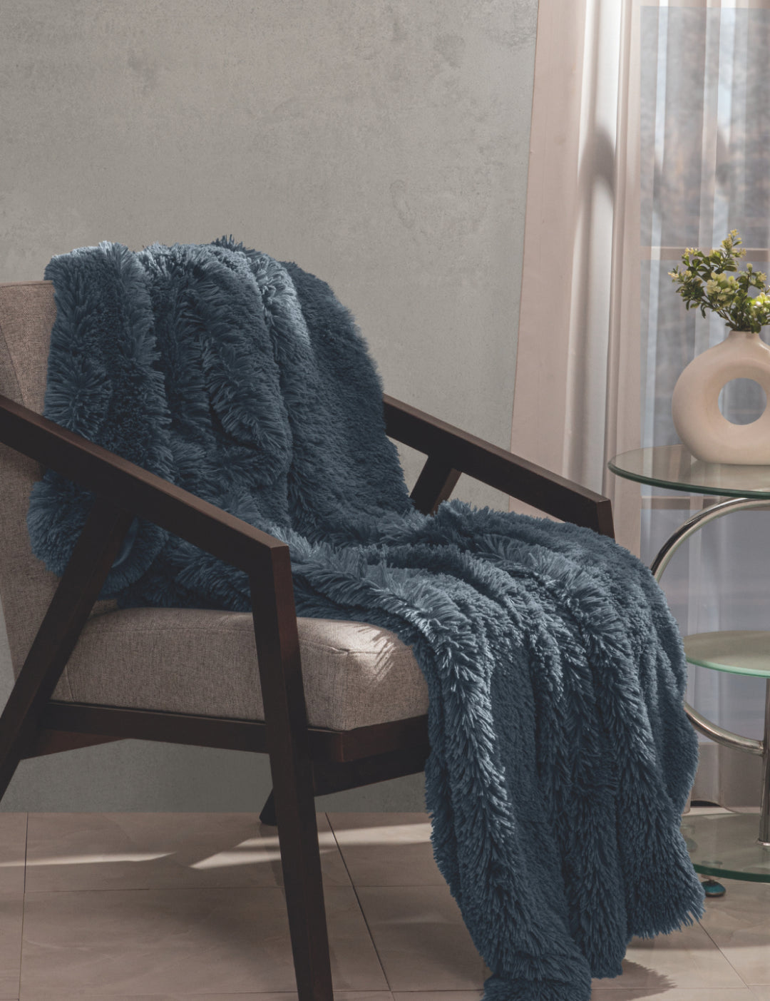 The Frazada de Lujo Bruselas de Intima Hogar, azul y con textura afelpada, decora un sillón junto a una mesa de vidrio, florero y planta, cerca de una ventana soleada con cortinas traslúcidas para máximo confort.