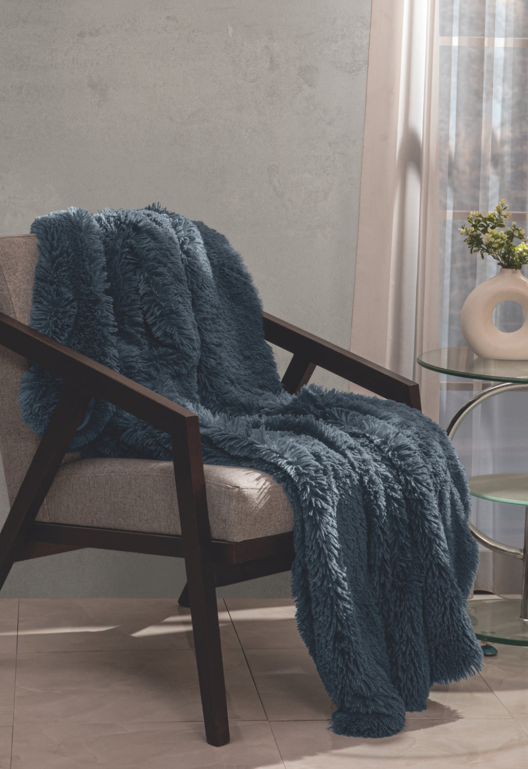 The Frazada de Lujo Bruselas de Intima Hogar, azul y con textura afelpada, decora un sillón junto a una mesa de vidrio, florero y planta, cerca de una ventana soleada con cortinas traslúcidas para máximo confort.