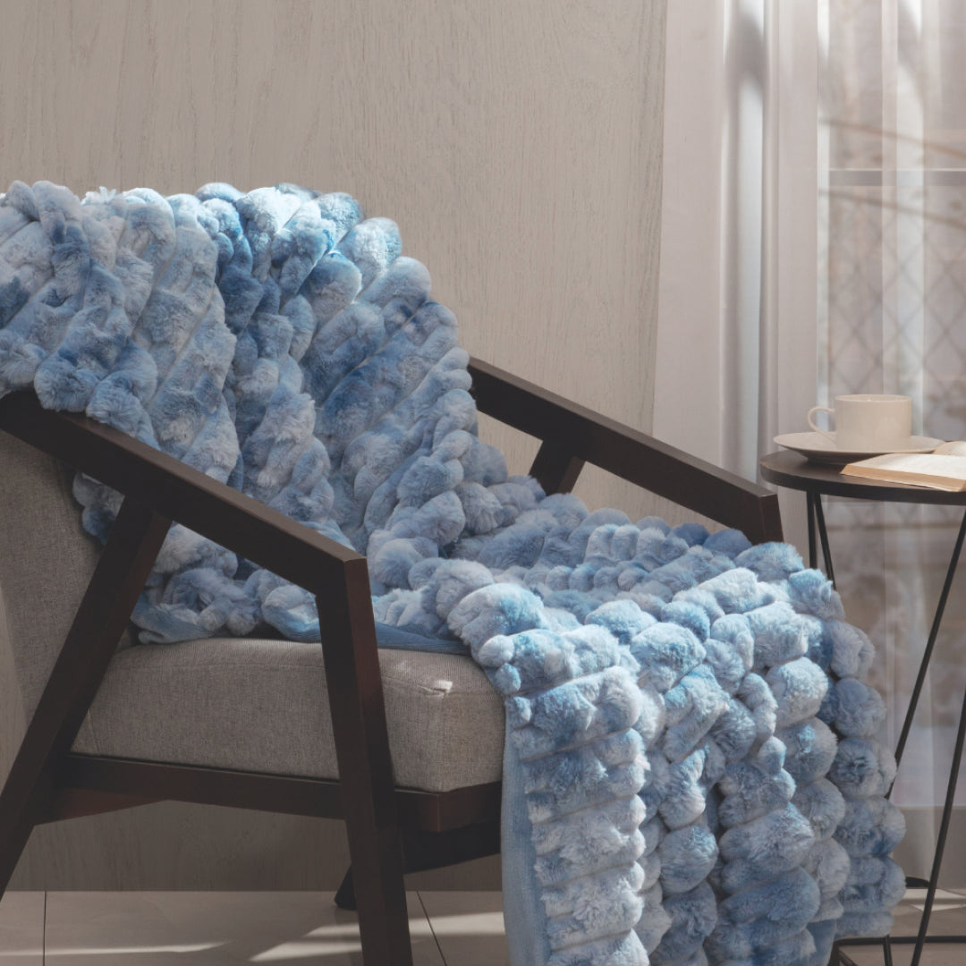 A luxurious Frazada de Lujo Polar by Intima Hogar in light blue is draped over a gray armchair beside a small round table with a cup and saucer, set in a sunlit room with sheer curtains.