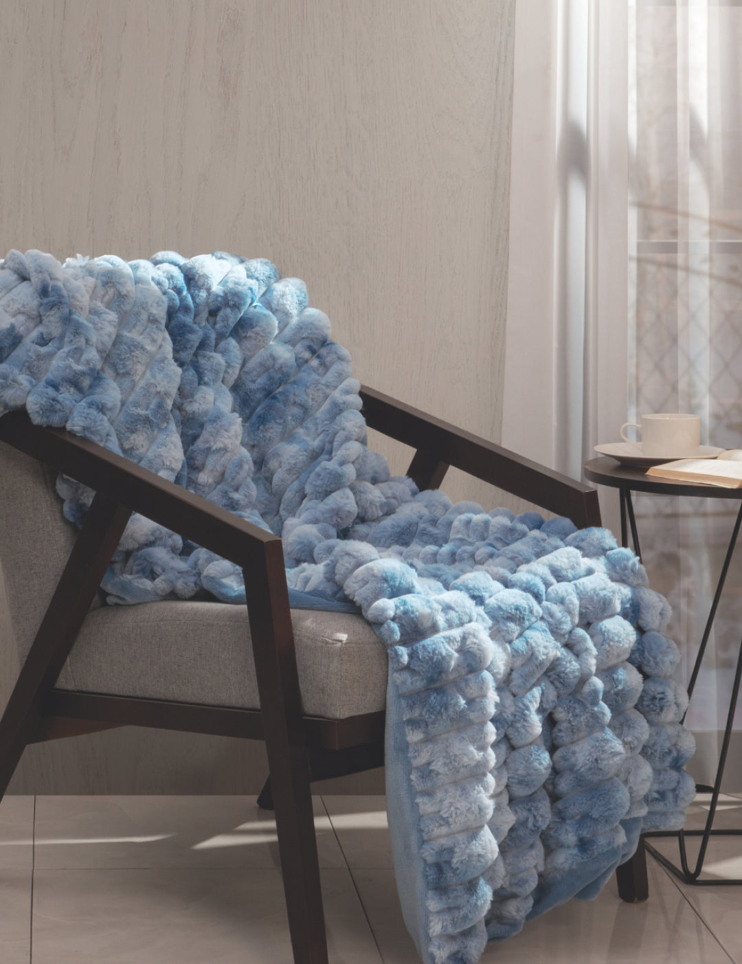 A luxurious Frazada de Lujo Polar by Intima Hogar in light blue is draped over a gray armchair beside a small round table with a cup and saucer, set in a sunlit room with sheer curtains.