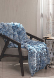 A luxurious Frazada de Lujo Polar by Intima Hogar in light blue is draped over a gray armchair beside a small round table with a cup and saucer, set in a sunlit room with sheer curtains.