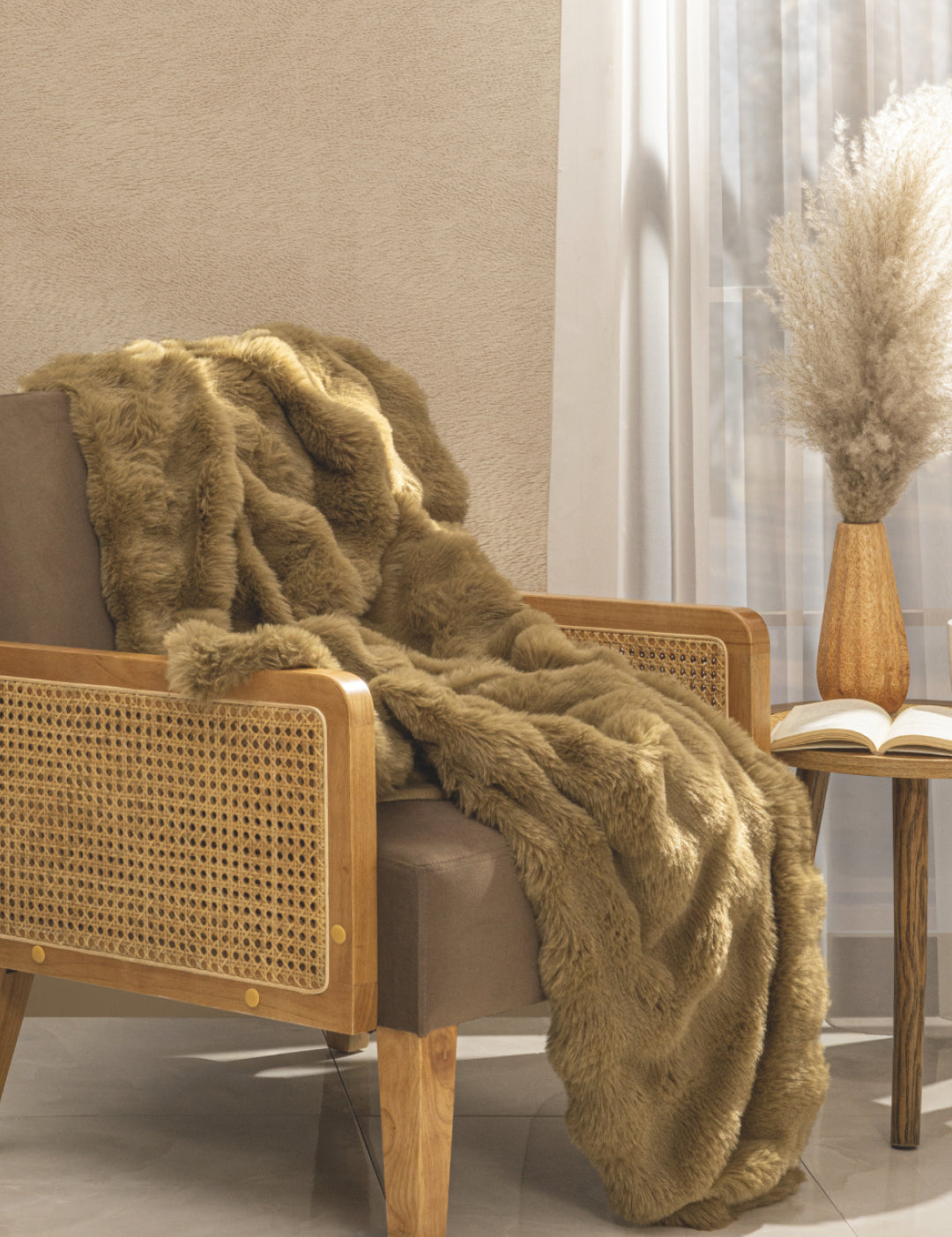 A cozy armchair with a wooden frame and rattan sides is draped with the Frazada de Lujo Quebec by Intima Hogar. Next to it, a round side table holds an open book and pampas grass by a sunlit window with white curtains.