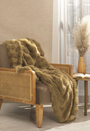 A cozy armchair with a wooden frame and rattan sides is draped with the Frazada de Lujo Quebec by Intima Hogar. Next to it, a round side table holds an open book and pampas grass by a sunlit window with white curtains.